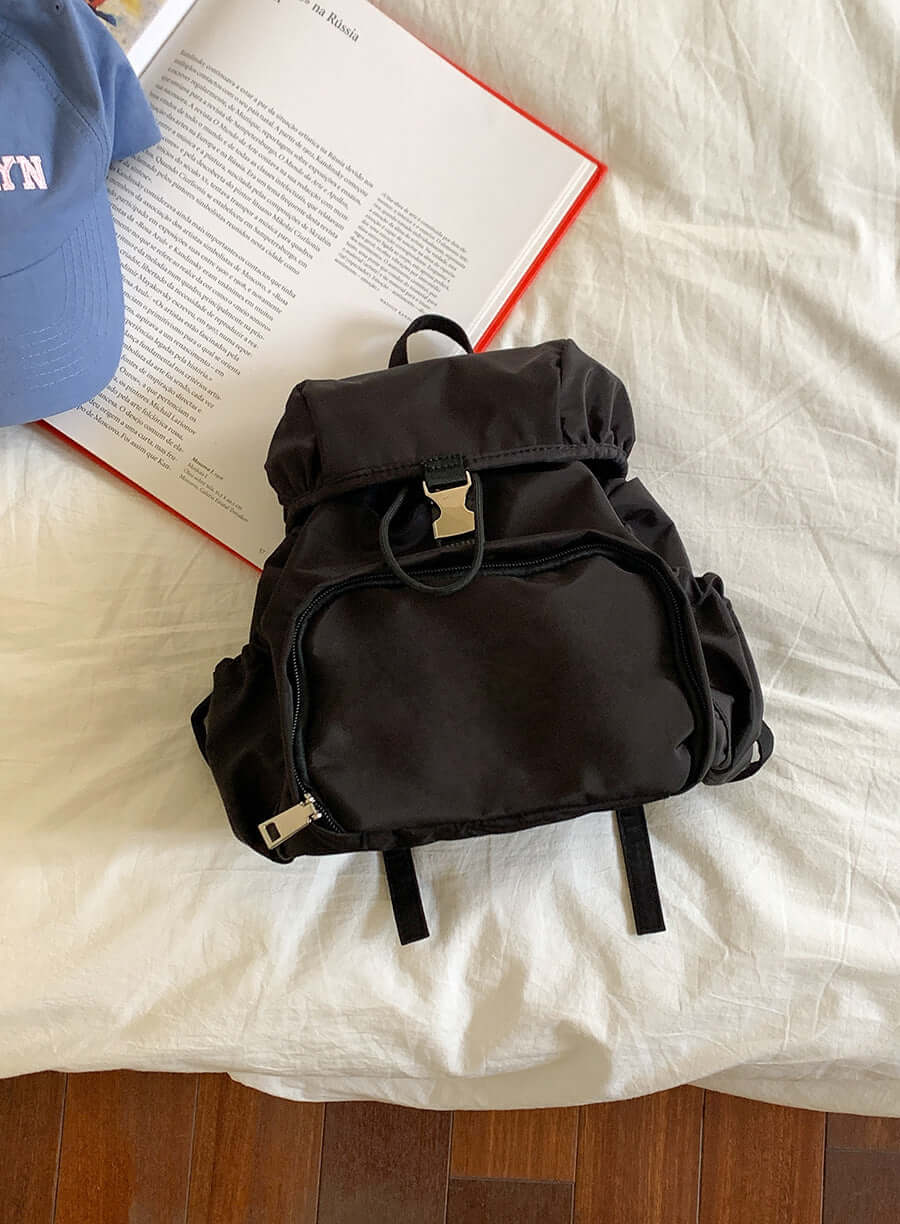 Black nylon backpack on a bed with a cap and book, perfect for casual outings and school use.