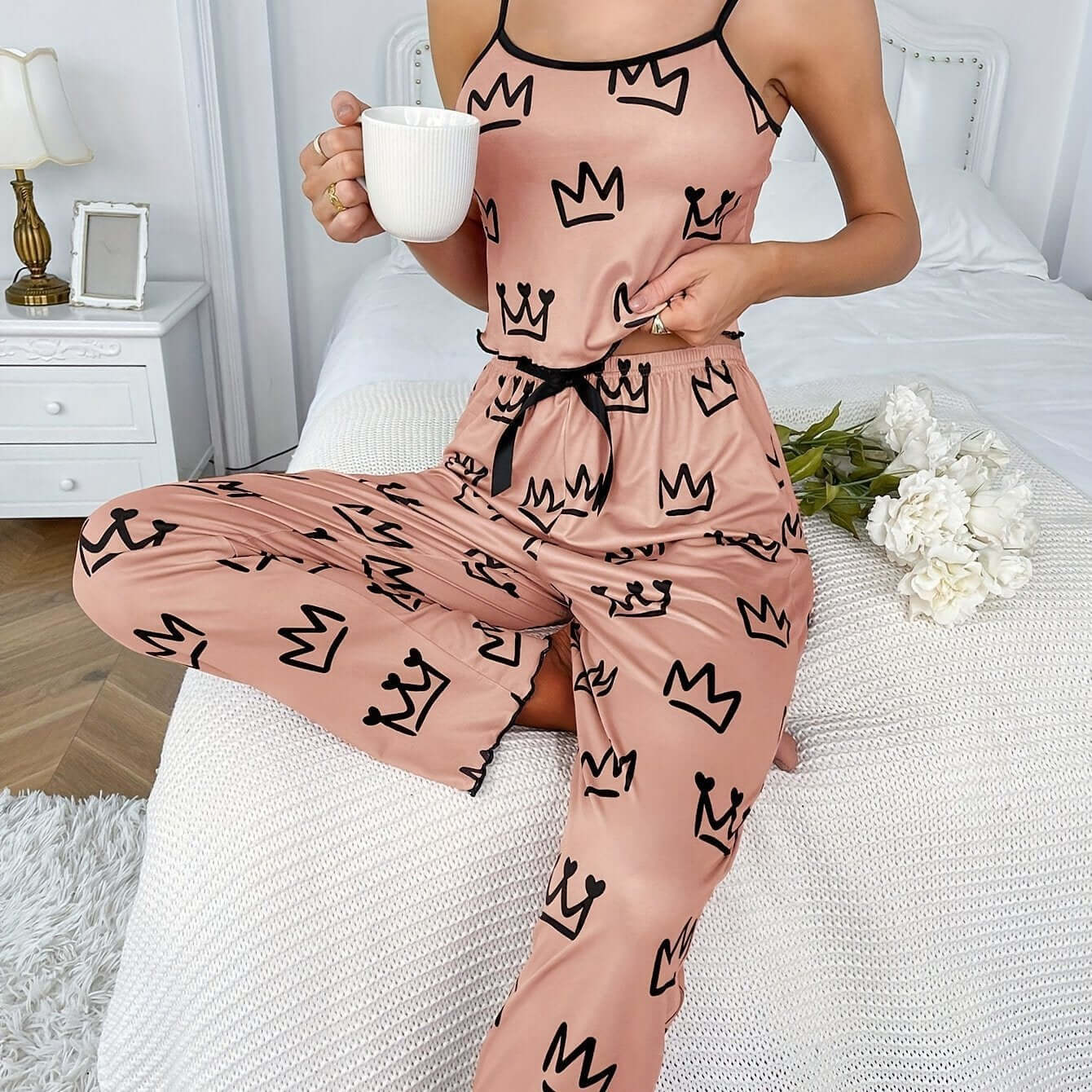 Stylish woman in pink two-piece milk silk suspender pajamas with crown pattern, enjoying coffee at home.