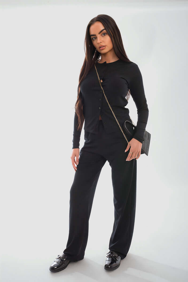 Model wearing a stylish black long sleeve button down top and trouser co-ord set, featuring a chic crossbody bag and sneakers.